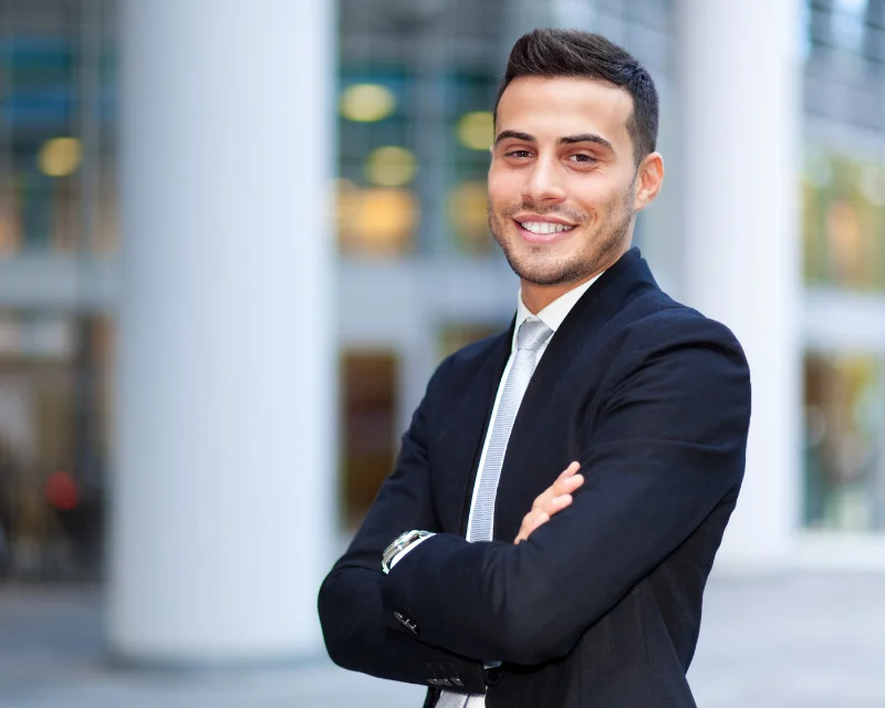 Smiling young man in dark suit and tie with arms crossed outside modern building